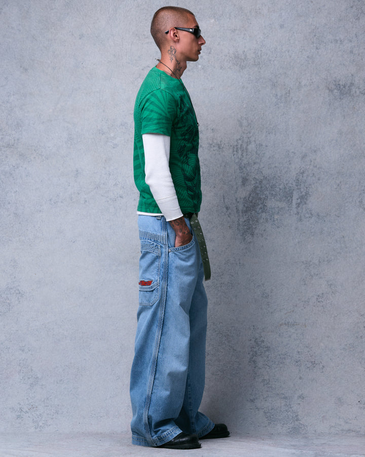 Model wearing light-wash wide-leg jeans, side view showing the leg piping and a red logo patch at the back pocket