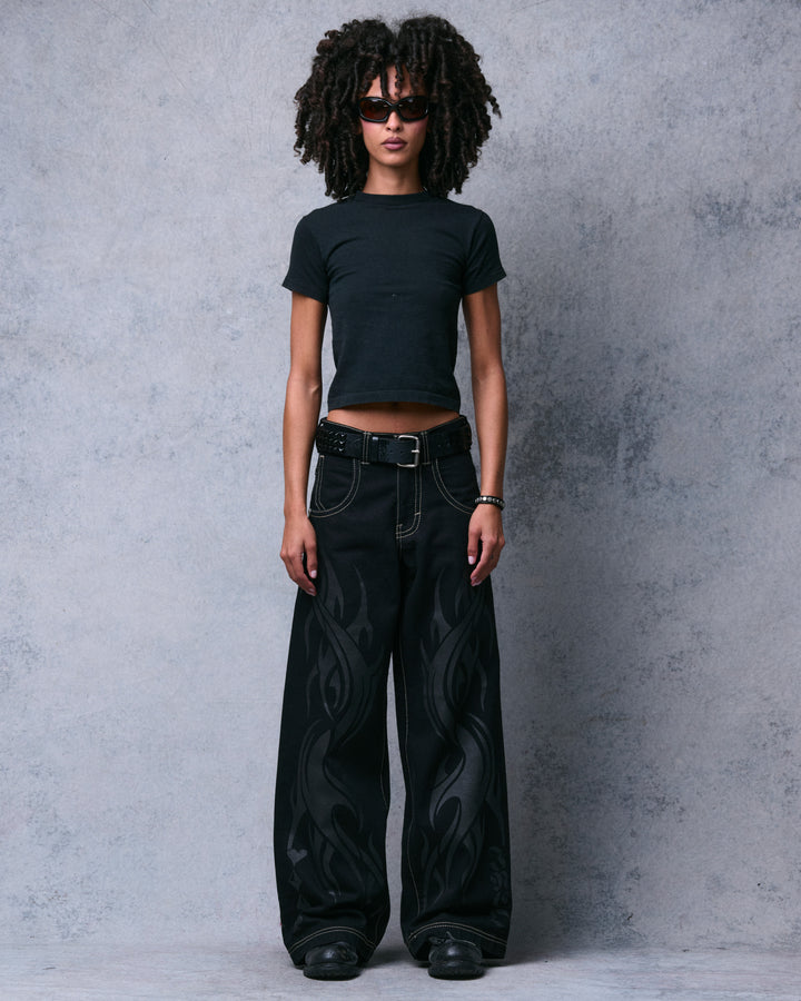 Model wearing black wide-leg jeans with tonal tribal flame print and a studded belt, standing straight, full front view