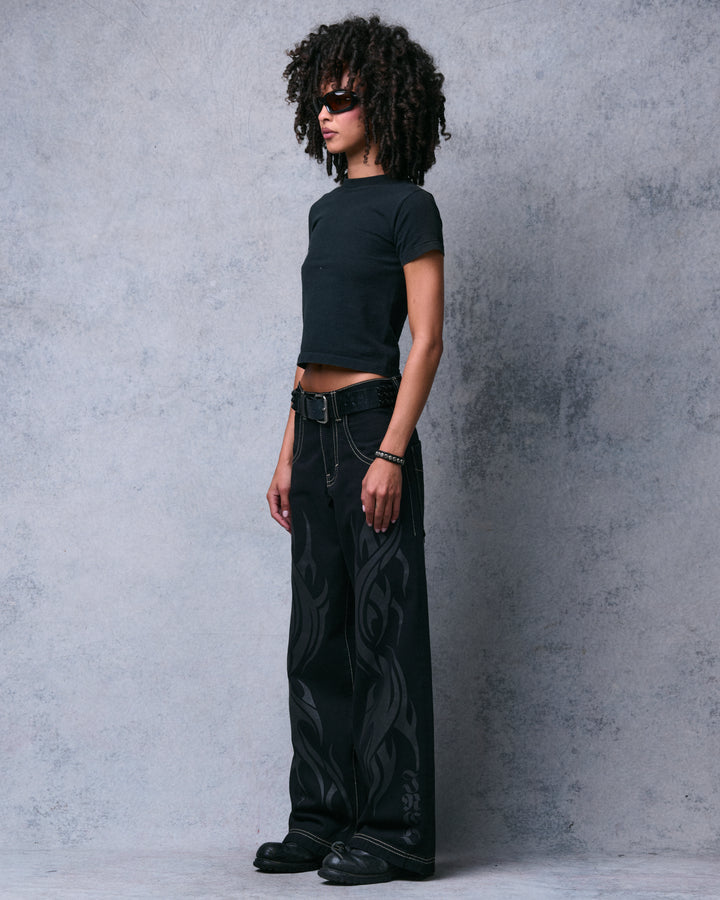 Model wearing black wide-leg jeans with tonal flame print and JNCO lettering near the hem, full side view