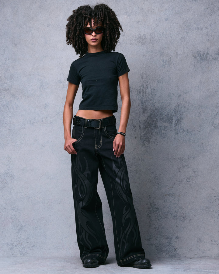 Model wearing black wide-leg jeans with tonal flame print, one hand at the front pocket, full front view at an angle