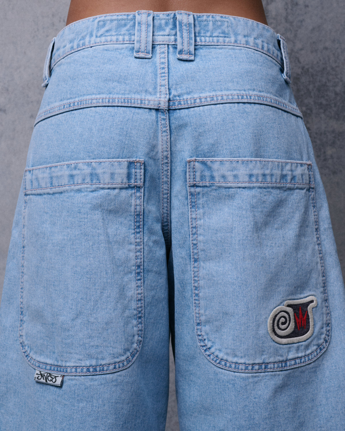 Back view of light-wash denim shorts with two wide patch pockets, one with an embroidered swirl crown patch