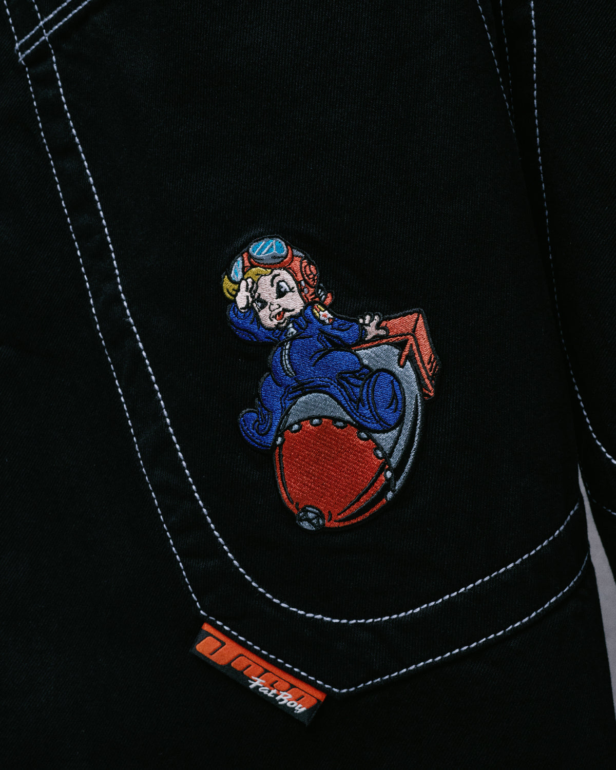 Close-up of black jeans' back pocket with embroidered cartoon pilot riding a bomb and an orange JNCO Fat Boy tab