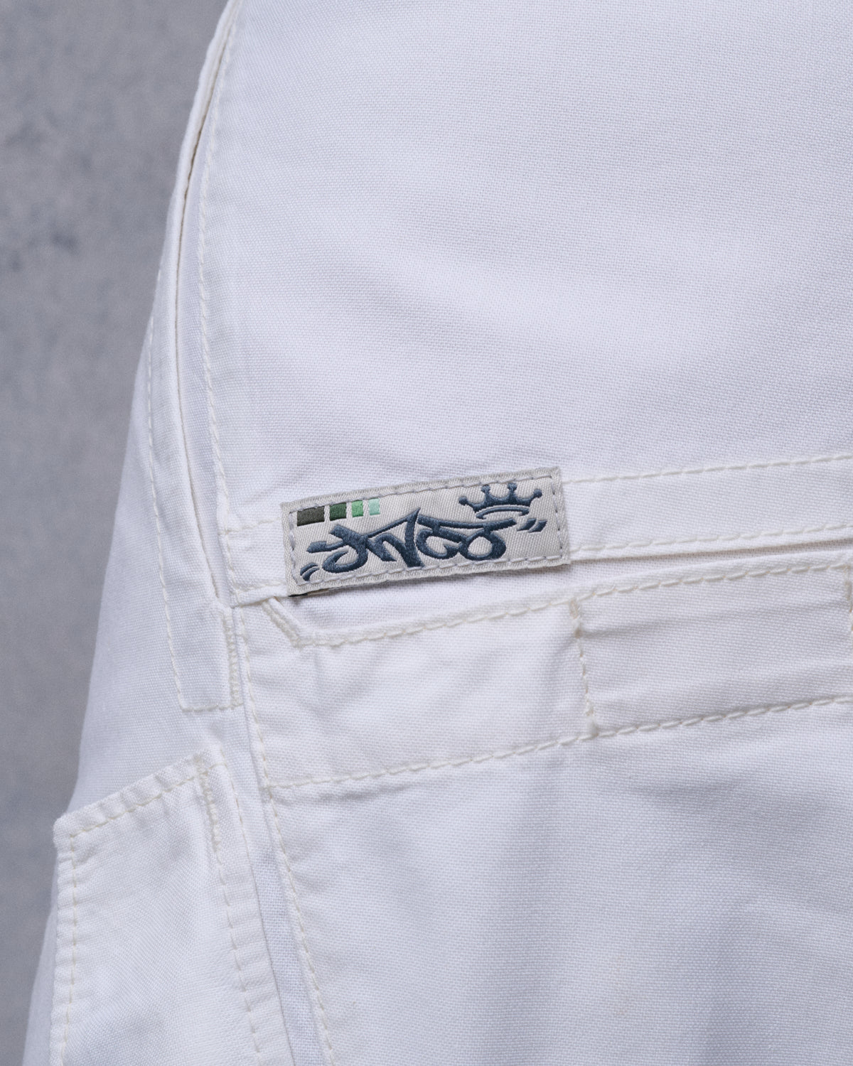 Close-up of a woven JNCO crown logo tab stitched above a welt pocket on off-white fabric