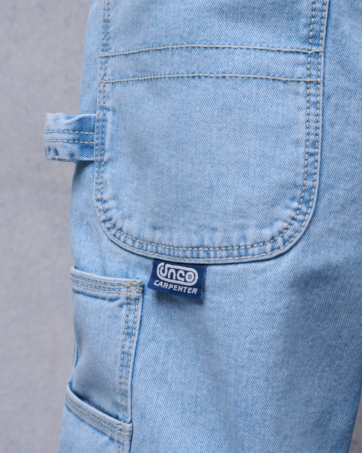 Close-up of light-wash denim shorts showing a rear pocket, hammer loop, side tool pocket and a navy JNCO Carpenter tab
