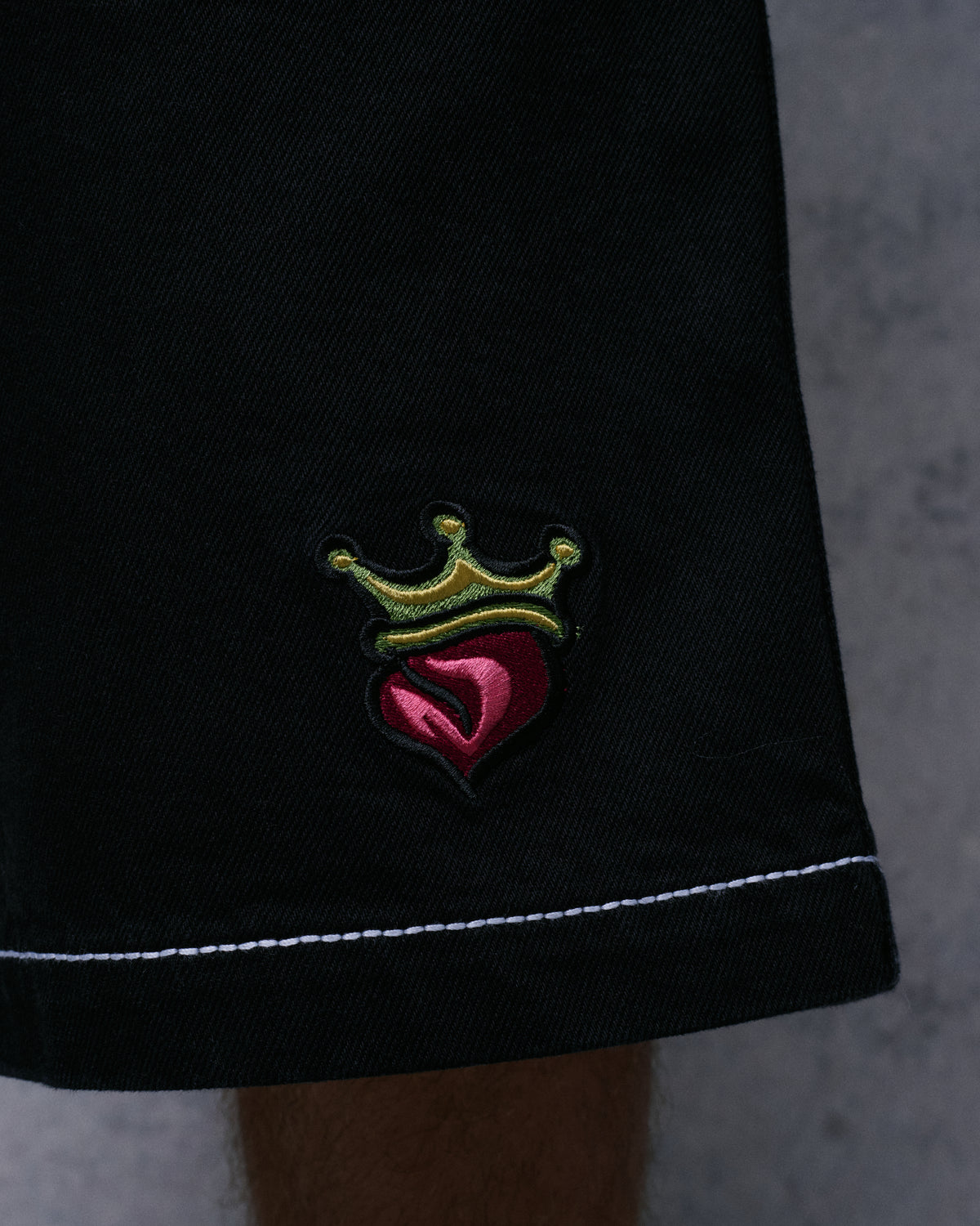 Close-up of the black denim shorts' leg hem with an embroidered crowned heart patch and white contrast stitching