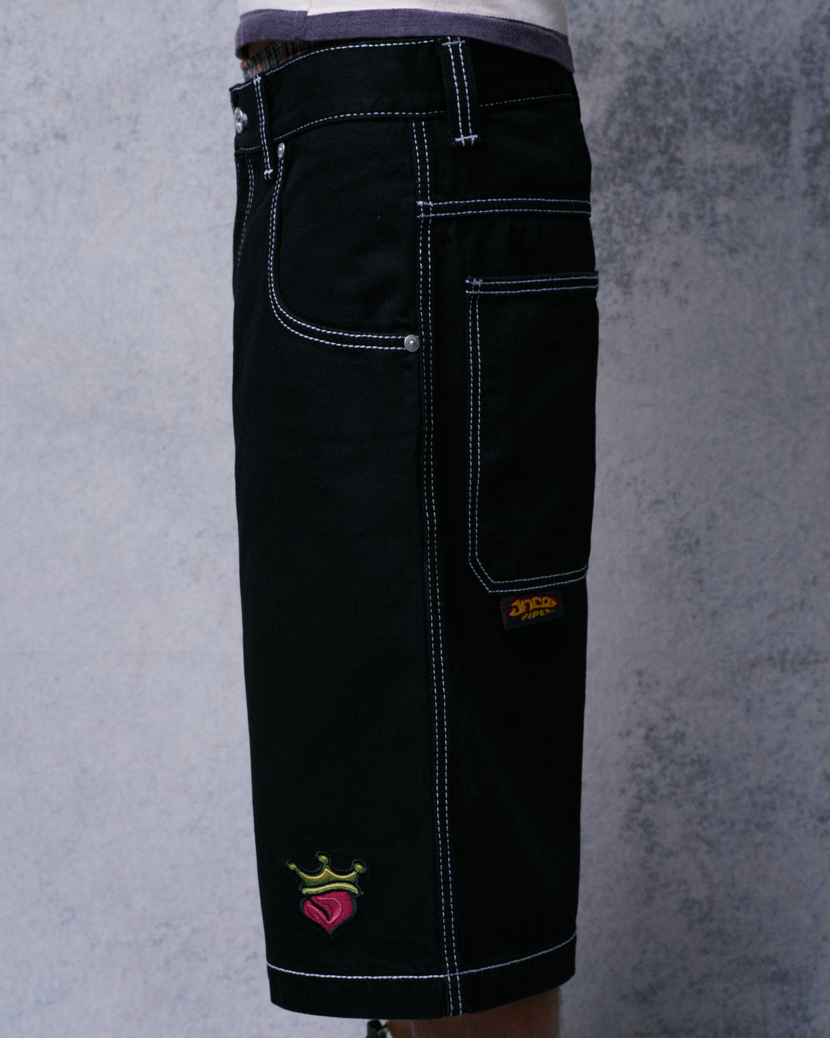 Side view of a model wearing the black denim shorts, showing white stitching, back pocket tab and crowned heart embroidery