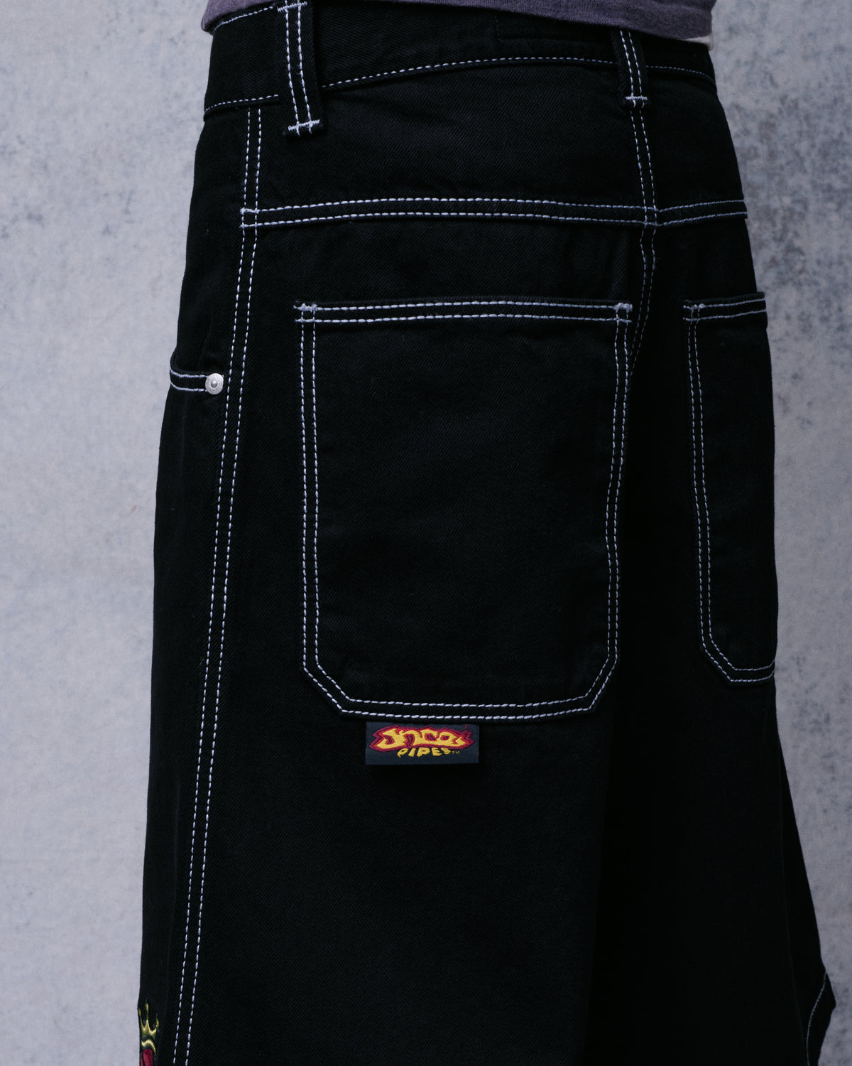 Angled back view of the black denim shorts showing a back pocket with white stitching and JNCO Pipes tab
