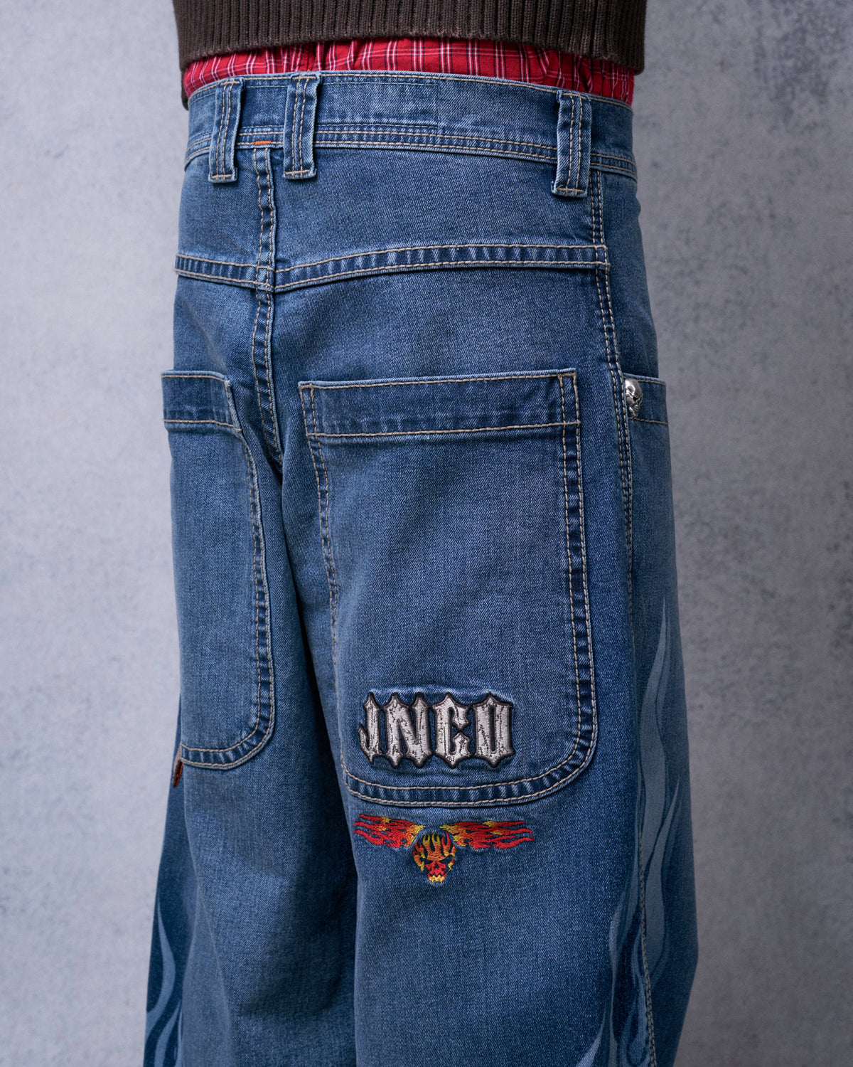 Back of the jeans on a model, showing a JNCO embroidered back pocket with a flaming skull below and tonal flames