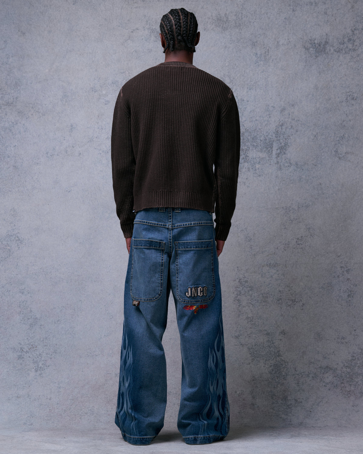 Model wearing wide-leg blue jeans, back view showing large back pockets, JNCO and red flame embroidery, and flame print