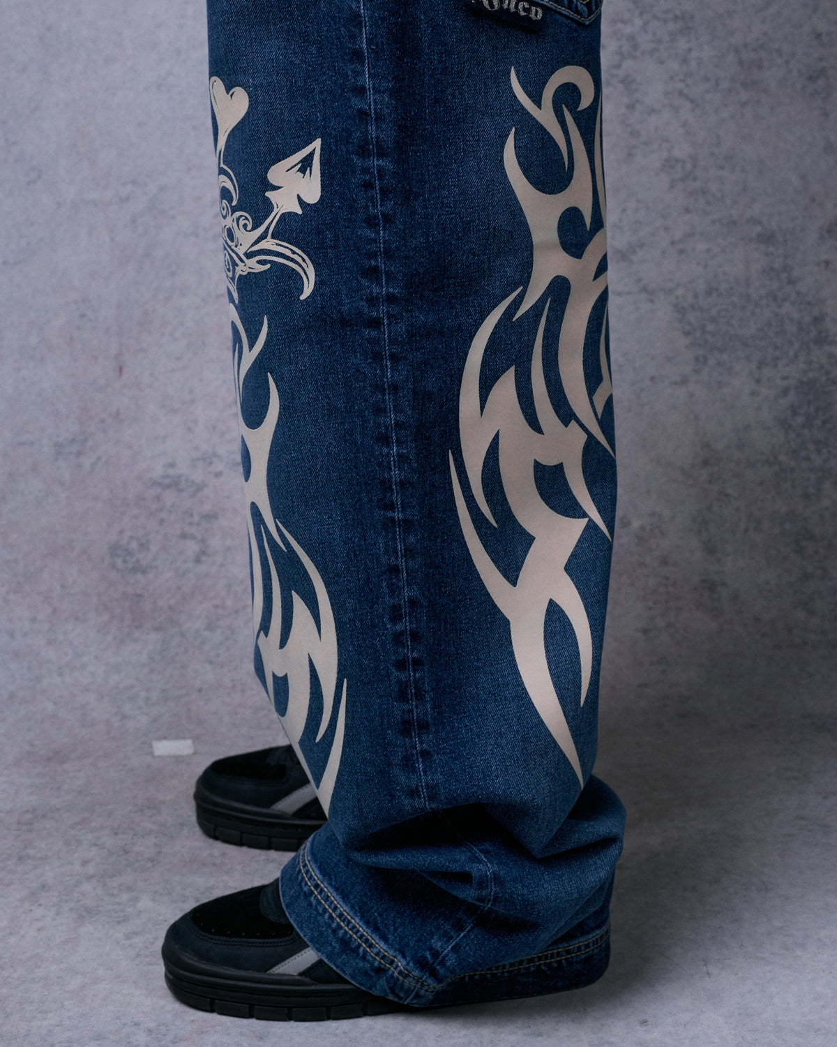 Side view of dark stone wide-leg jeans with cream tribal graphics, hems bunched over black sneakers