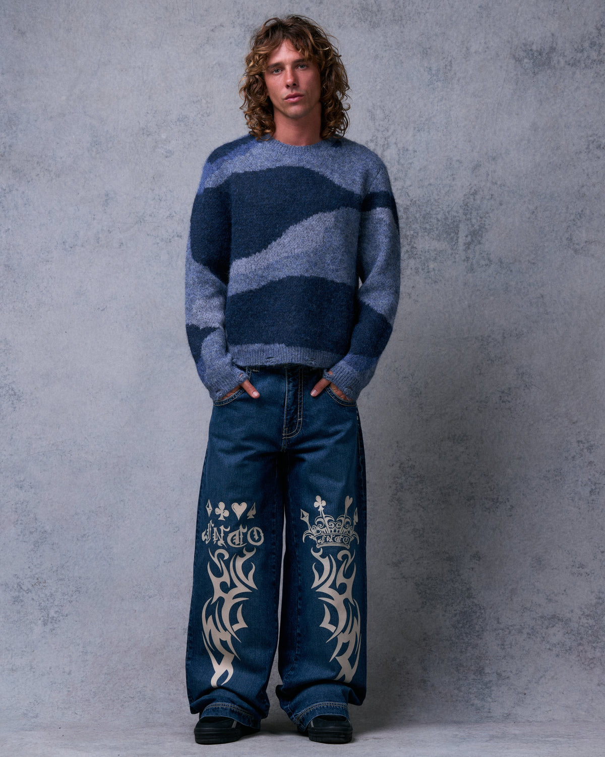 Model wearing dark stone wide-leg jeans with cream tribal, crown and card suit graphics on the front legs, front view