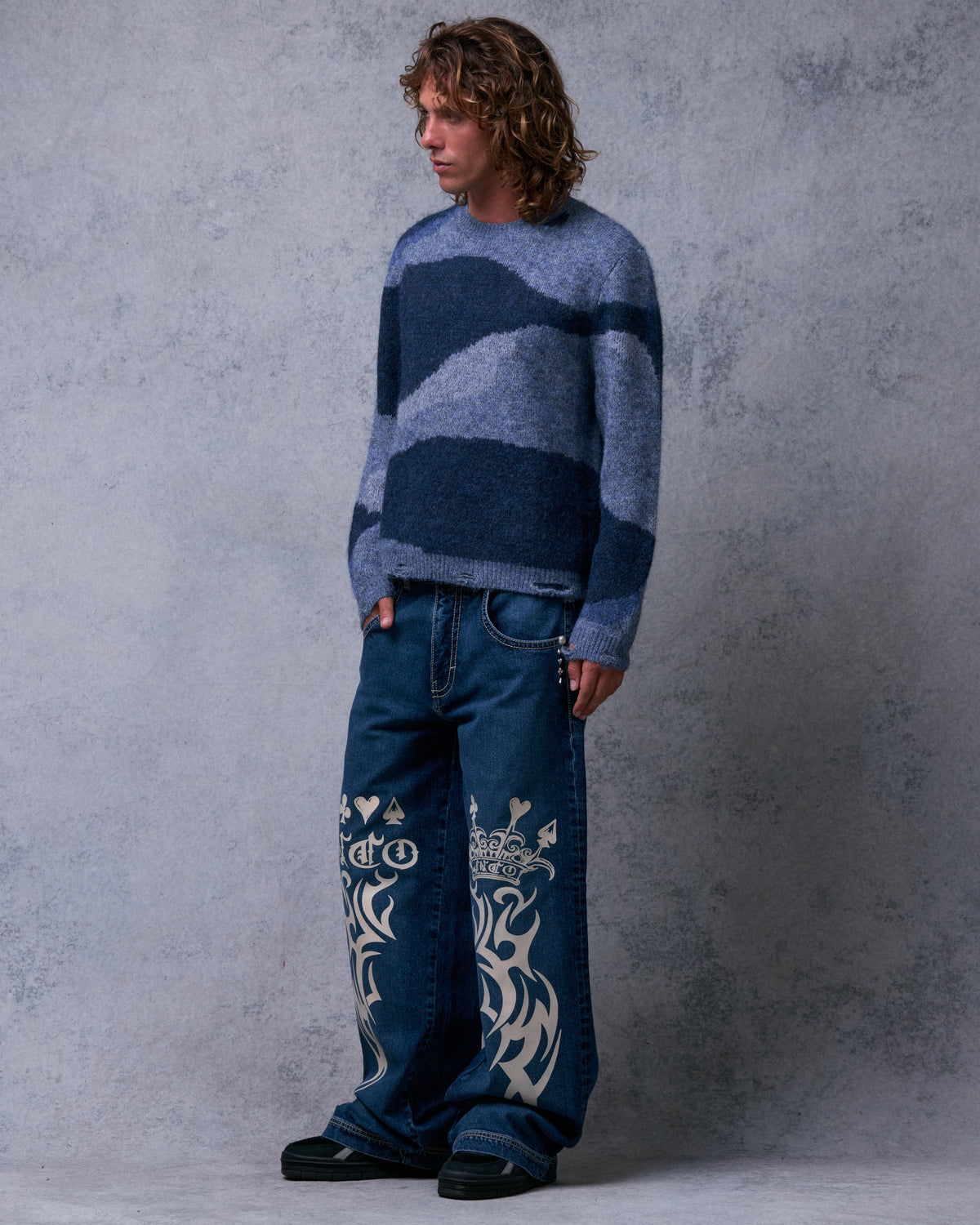 Model wearing the dark stone wide-leg jeans, three-quarter front view showing the crown and tribal leg graphics