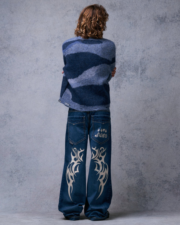Model wearing the dark stone wide-leg jeans from behind, showing tribal graphics on the back legs and a logo on one pocket