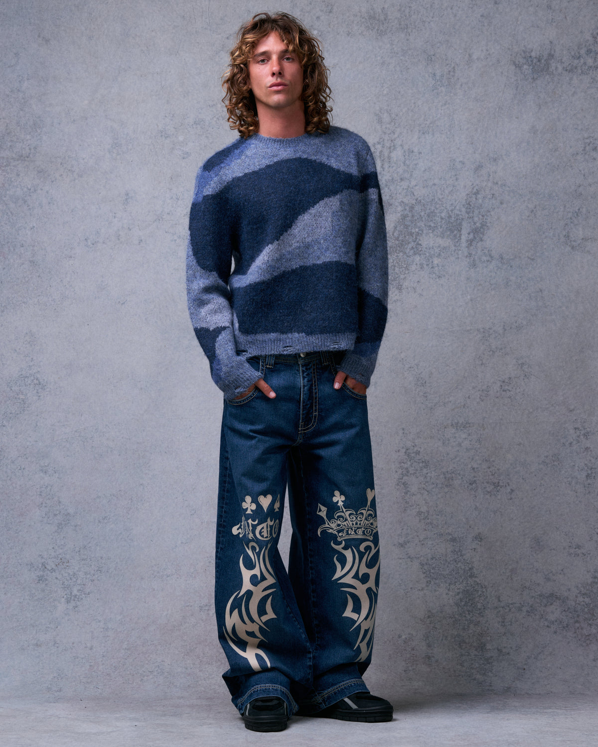 Model wearing dark stone wide-leg jeans with cream tribal, crown and card suit graphics on both legs, front view