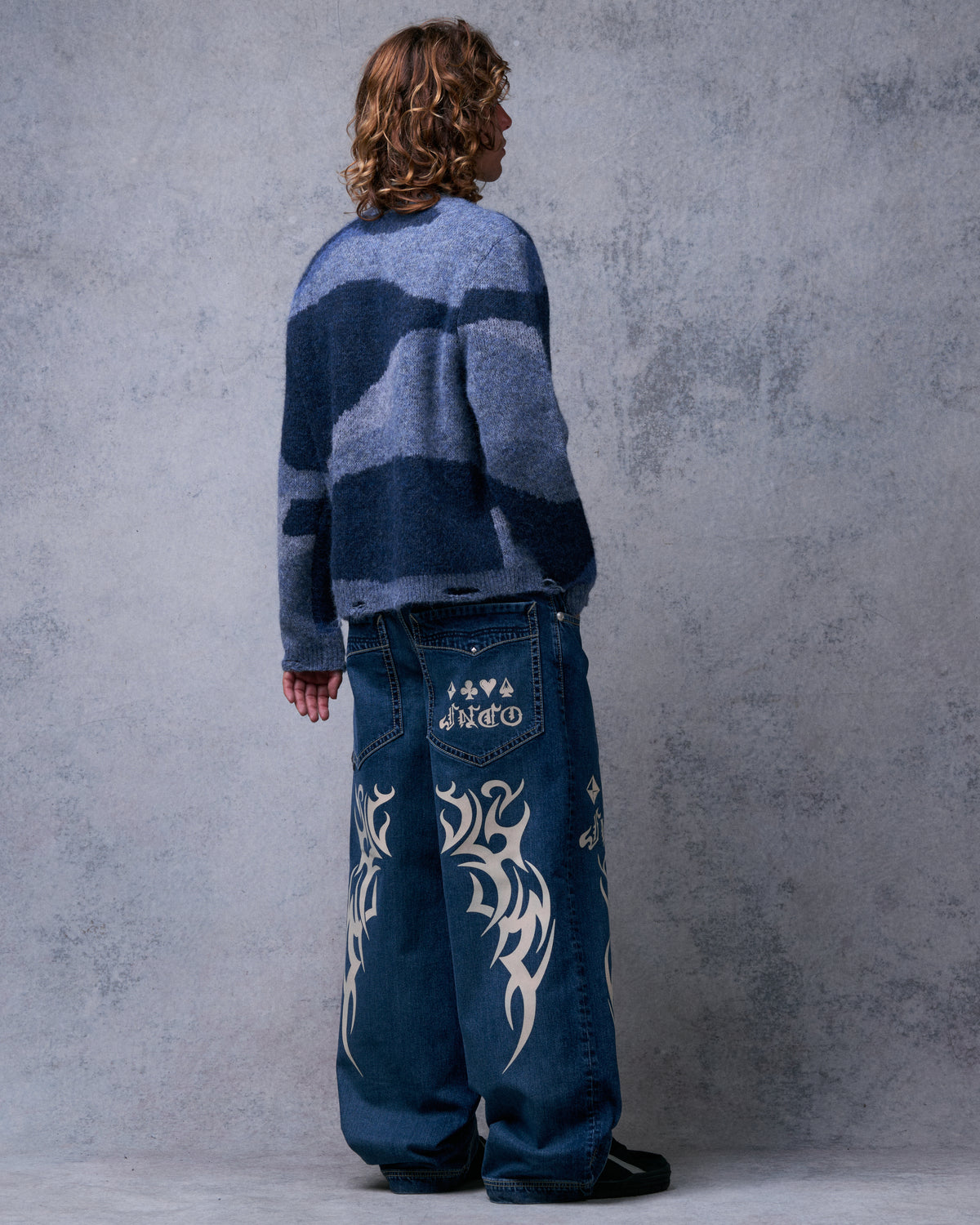 Model wearing the dark stone wide-leg jeans, three-quarter back view with card suit logo pocket and tribal back-leg graphics
