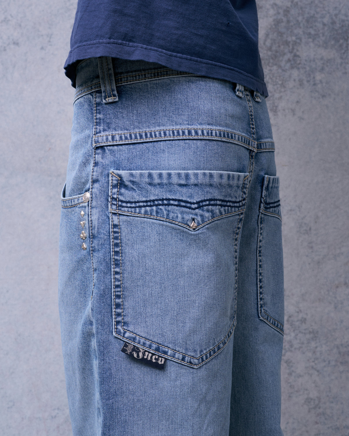 Back pocket of light blue wide-leg jeans with a spade stud and a black JNCO tab, rear three-quarter view