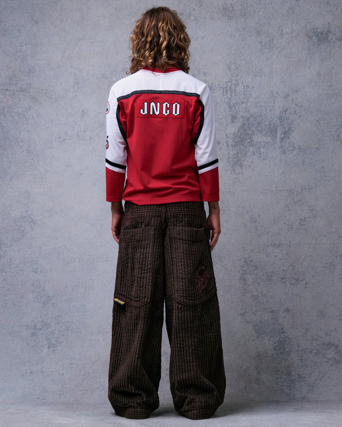 Model wearing brown wide-leg corduroy pants with a red jersey, back view showing large back pockets and crown J patch