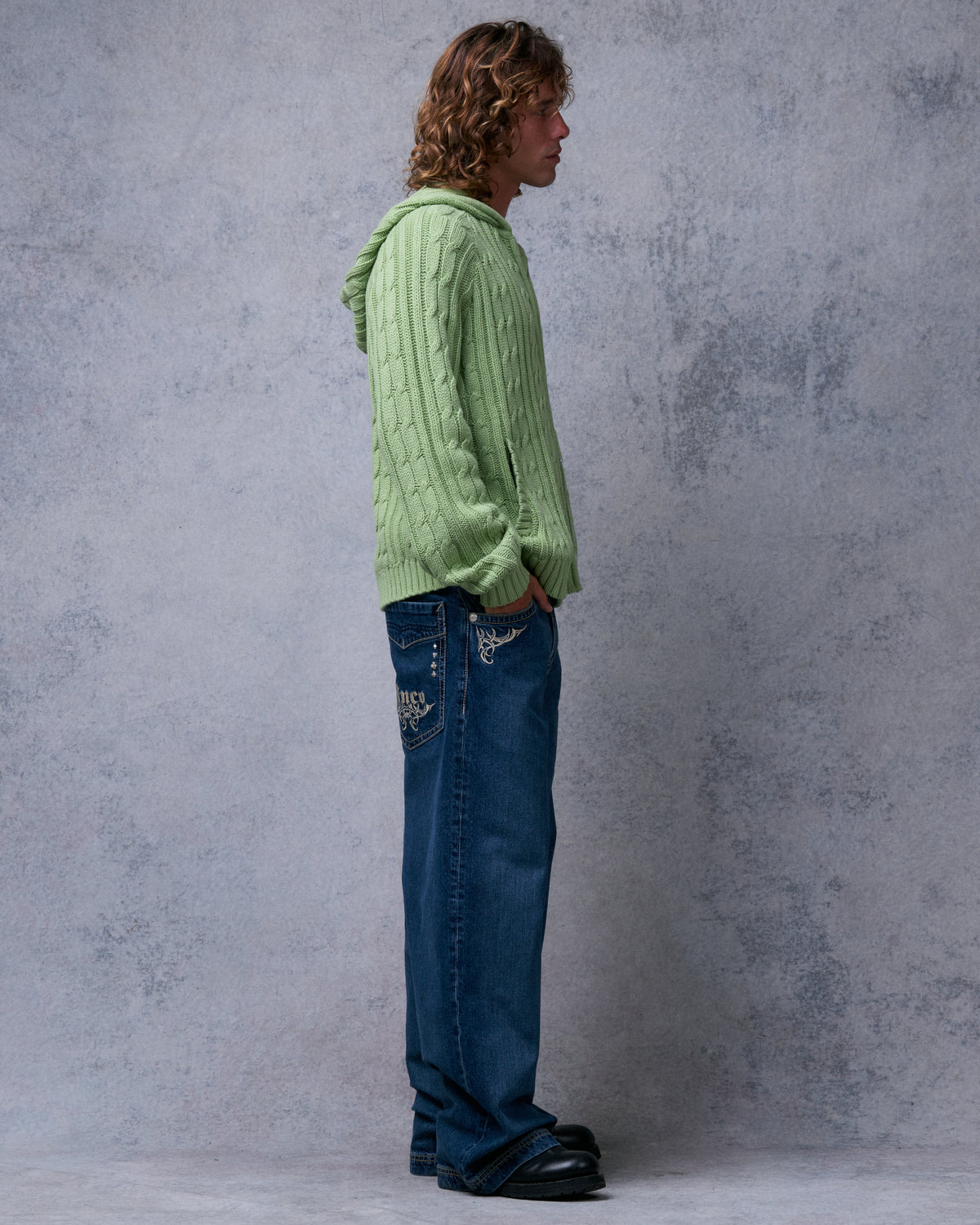 Model wearing dark stone wide-leg jeans with a green cable-knit hoodie, side view showing tribal pocket embroidery