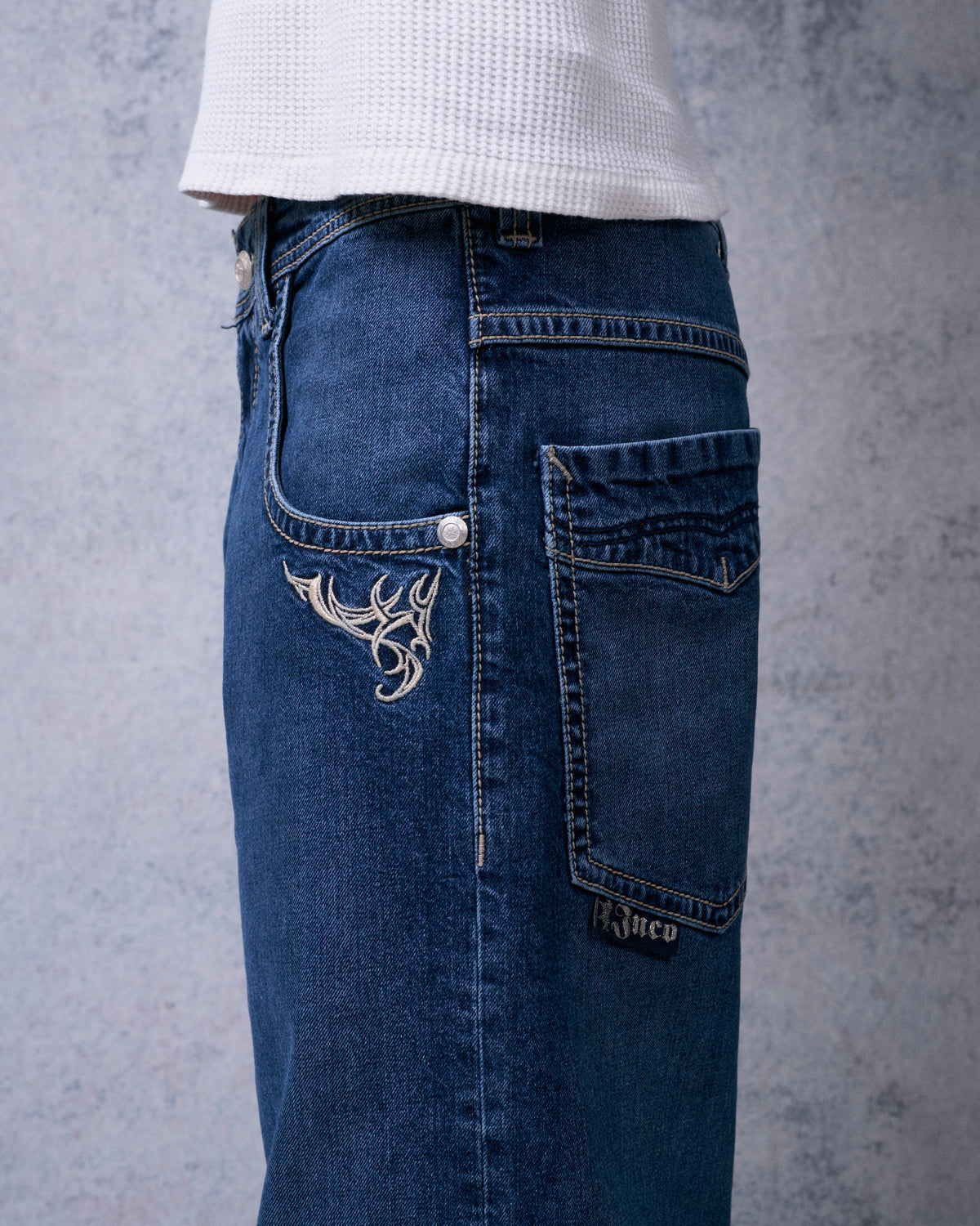 Side close-up of the dark stone jeans showing front tribal embroidery, back pocket and a small JNCO label tab