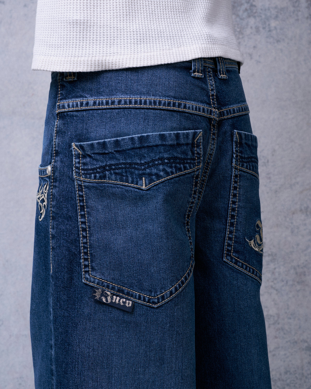 Back of dark stone wash jeans from an angle, showing contrast-stitched back pockets and a small JNCO tab label