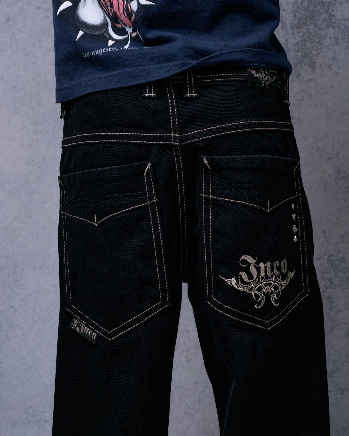Model wearing the black wide-leg jeans, back view with Jnco tribal embroidery and card-suit rivets on one pocket