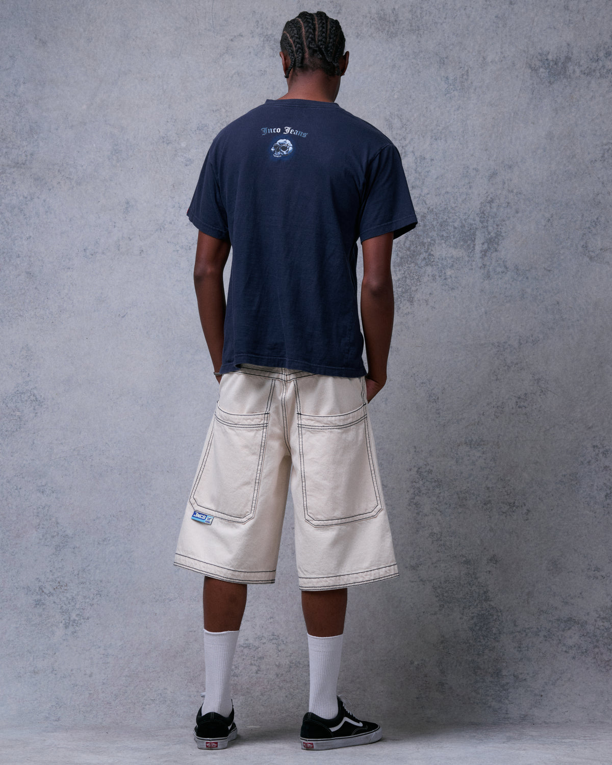 Back view of model wearing natural off-white denim shorts, showing wide double-stitched back pockets and a blue label