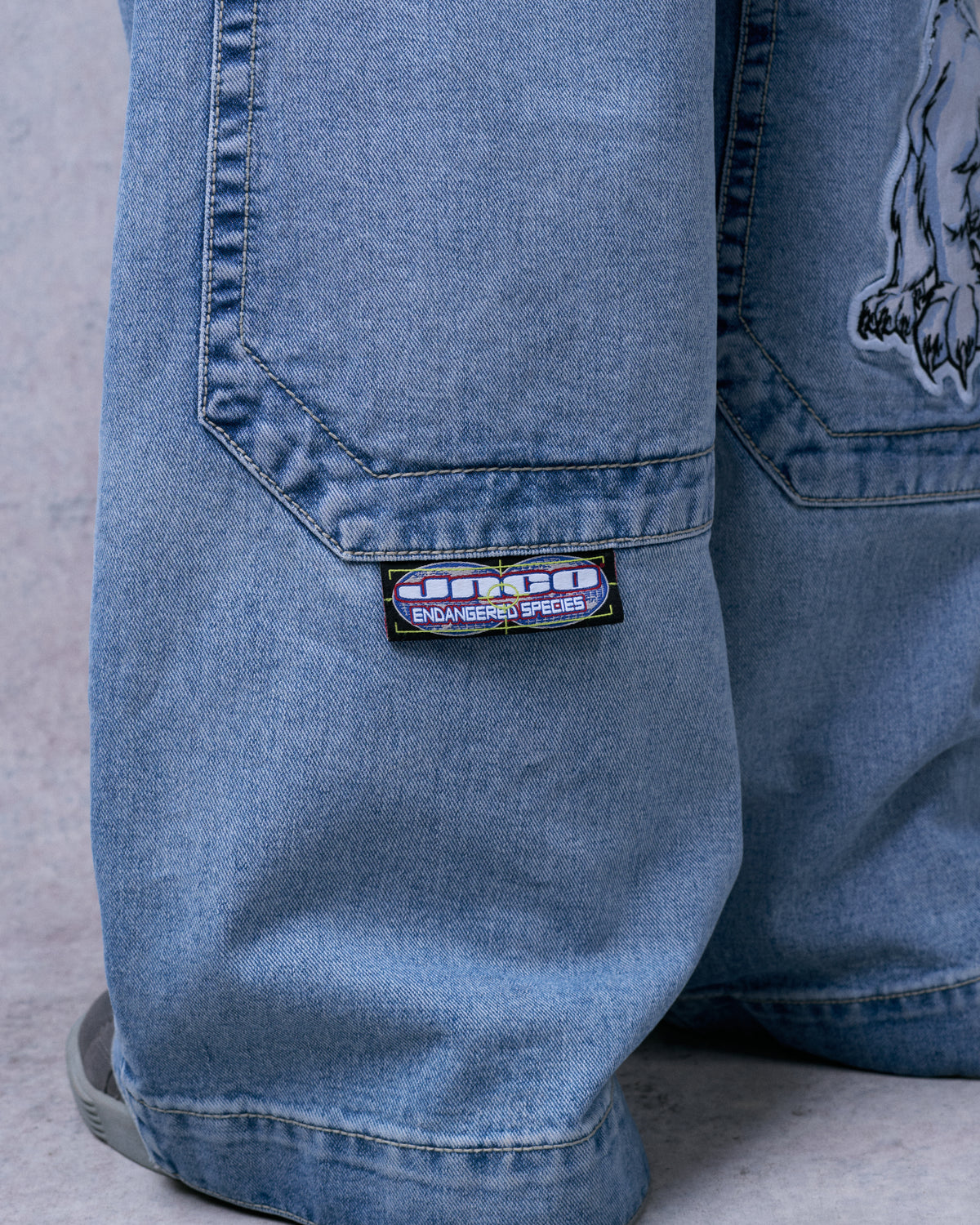 Close-up of light stone wash wide-leg jeans, back of the leg with a JNCO Endangered Species woven tab below the pocket
