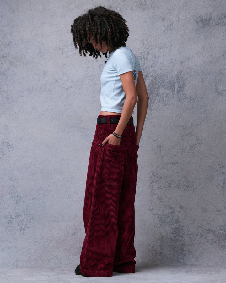 Model wearing burgundy wide-leg corduroy pants, side view with hand in front pocket and back pocket visible